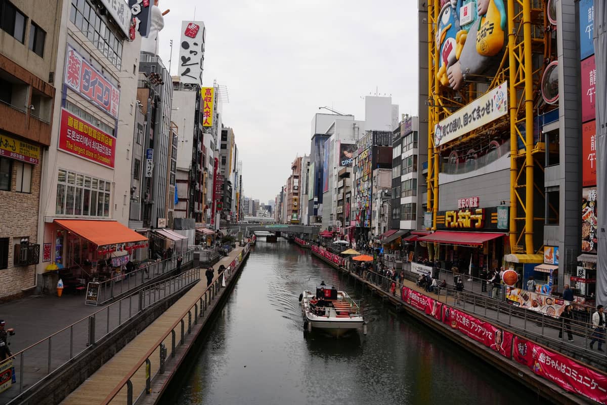 Osaka's Dotonbori canal with vibrant storefronts, large colorful signs, and a boat cruising down the water, bustling food and dining district atmosphere.