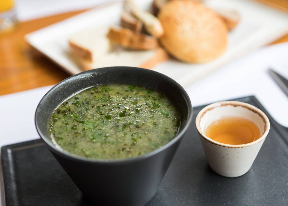 Eine Schüssel grüne Suppe mit Kräutern neben einer kleinen Tasse, mit Brot im Hintergrund auf einem Tablett.