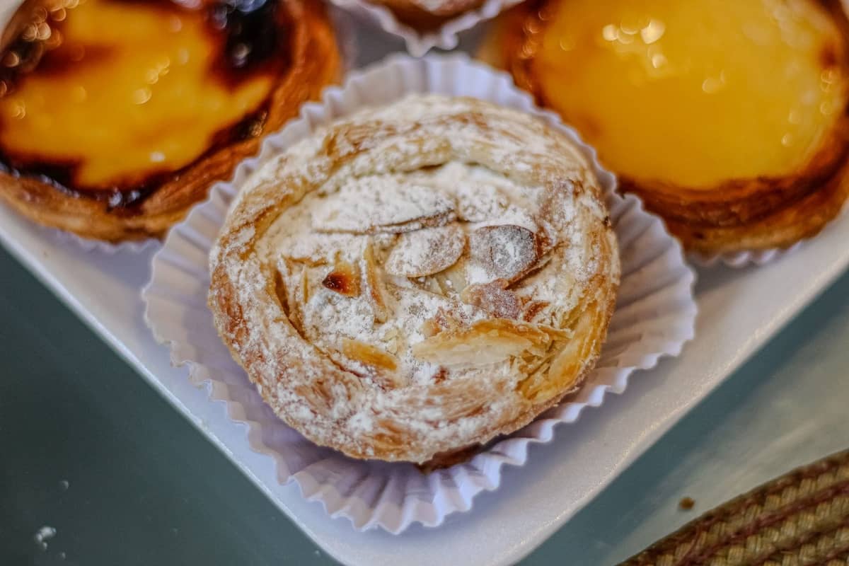 Nahaufnahme eines Jesuíta-Gebäcks, bestreut mit Puderzucker und Mandelblättchen, mit Pastéis de Nata in einer Bäckereibox daneben.