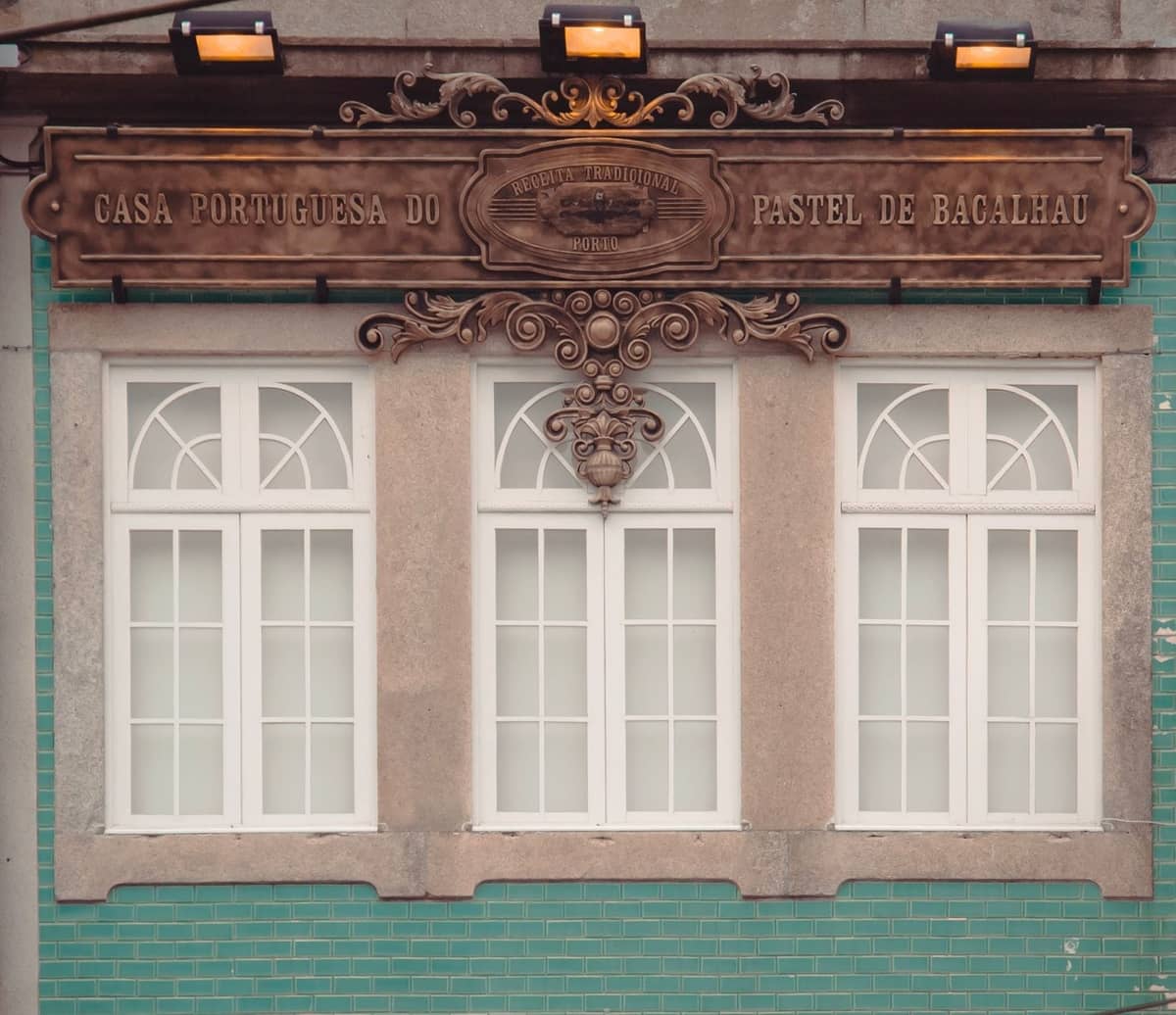 Außenansicht der Casa Portuguesa do Pastel de Bacalhau, einem Spezialgeschäft für Salzfisch-Snacks in Porto, mit seinem verzierten Schild über der grün gefliesten Fassade.