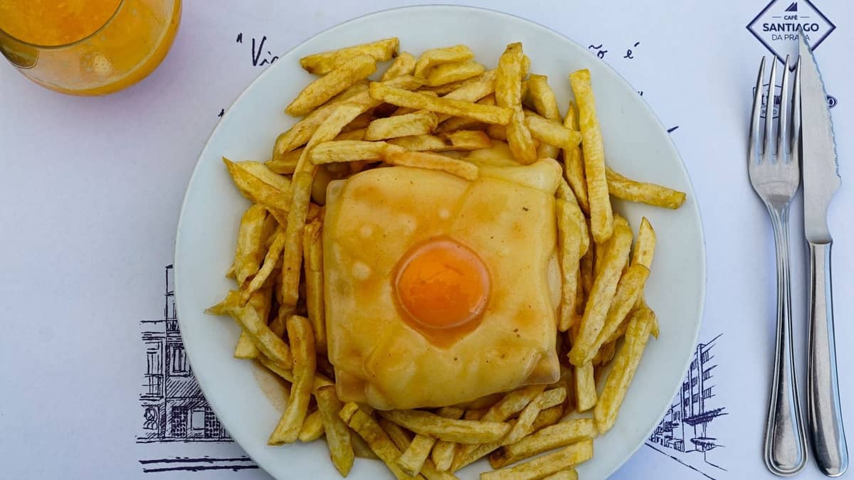 Ein Teller Francesinha, belegt mit einem Spiegelei und umgeben von Pommes frites, serviert auf einem weißen Teller mit Besteck und einem Orangengetränk.