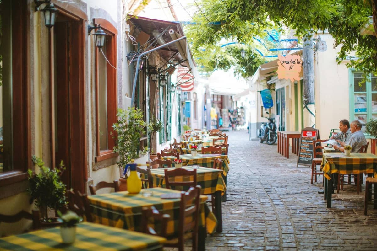Charming outdoor restaurant seating with green-checked tablecloths along a stone-paved alley in a sunlit Rhodes old town street.