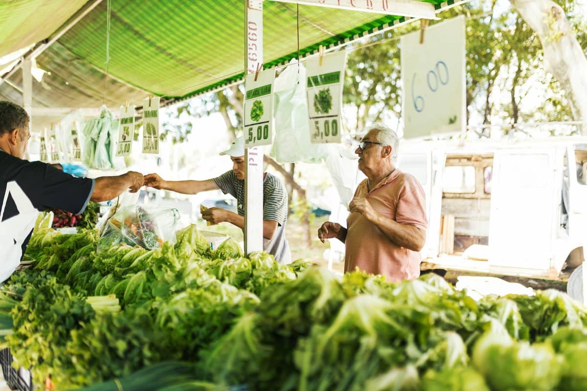 Local outdoor market stand in Rhodes with fresh green vegetables and herbs, vendors and customers trading under a shaded awning.