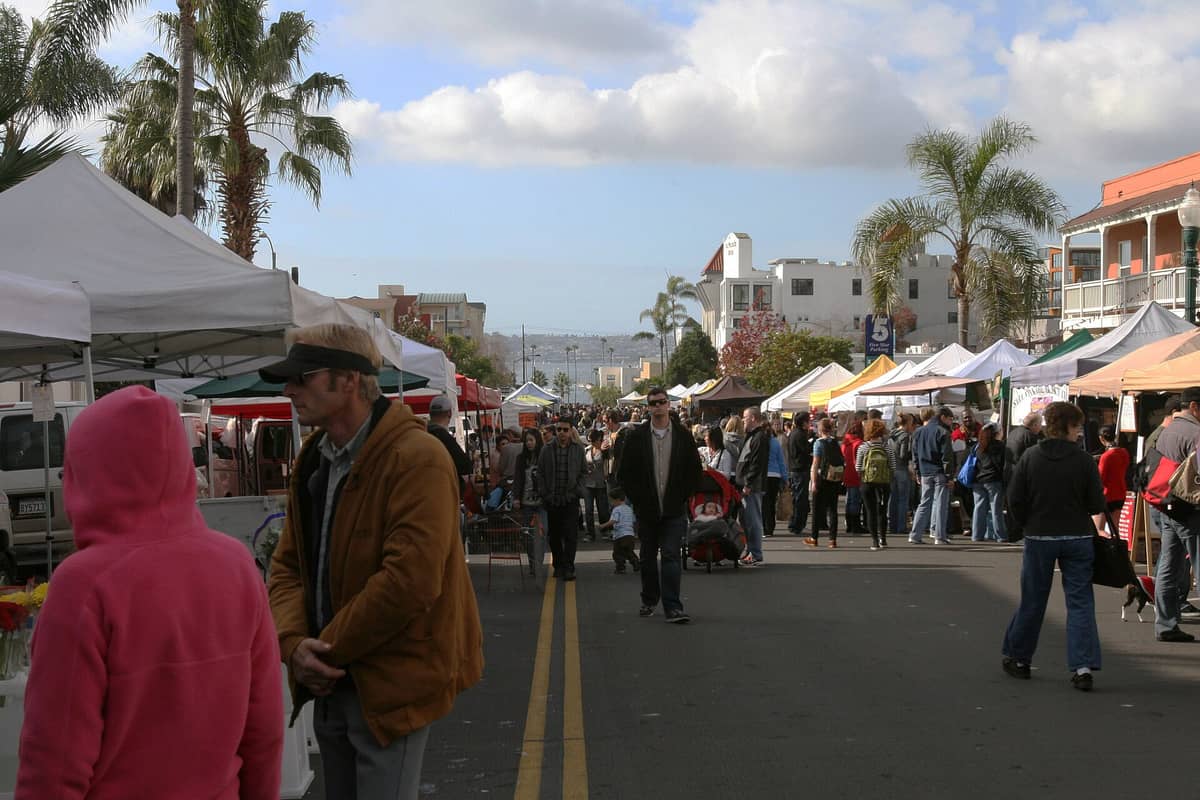 Menschenmassen beim Einkaufen an Außenständen auf einem großen Bauernmarkt, mit Palmen und Stadtgebäuden im Hintergrund.