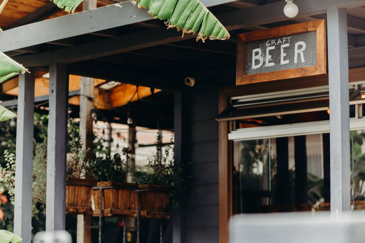 Außenansicht einer Bar oder eines Cafés mit einem Holzschild mit der Aufschrift 'Craft Beer' und sichtbarem Grün und Pflanzen rund um den Eingang.