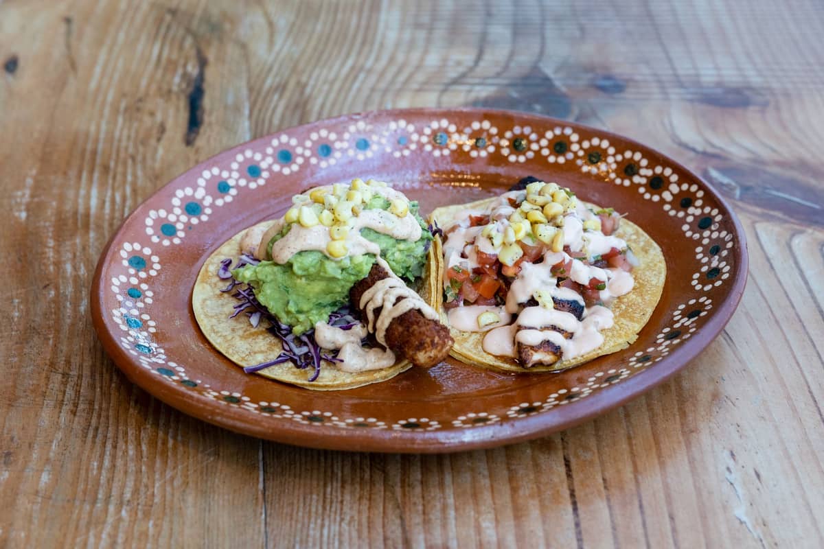 Zwei frittierte Fisch-Tacos mit Guacamole, Kohl, Mais, Pico de Gallo und Sauce auf einem braunen Dekorteller auf rustikalem Holz.