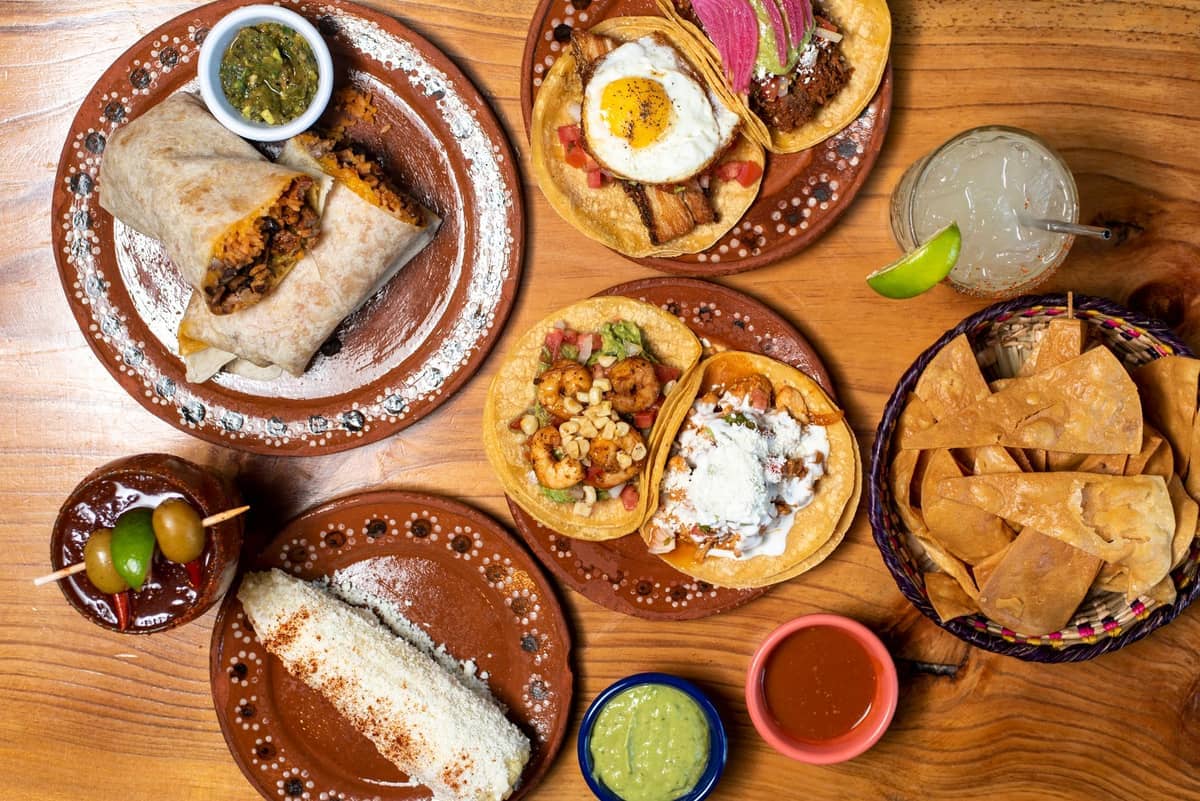 Una colorida variedad de platos mexicanos que incluye tacos, burrito, elote, tostadas y cócteles sobre una mesa de madera en San Francisco.