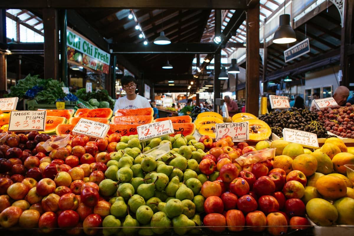 Indoor farmers market produce stands with apples, pears, and citrus fruits arranged in rows under wooden beams.