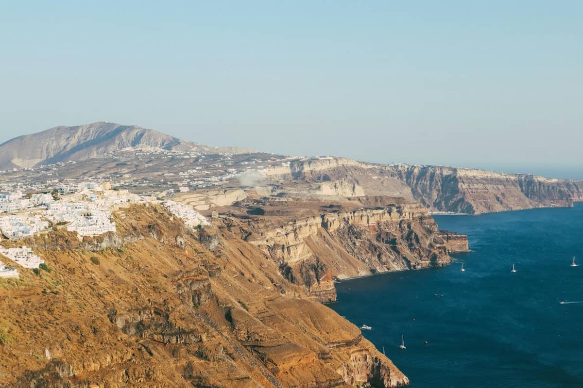 Aerial view of Santorini’s rugged volcanic cliffs with whitewashed towns perched on the edge and deep blue sea below, on a clear day.