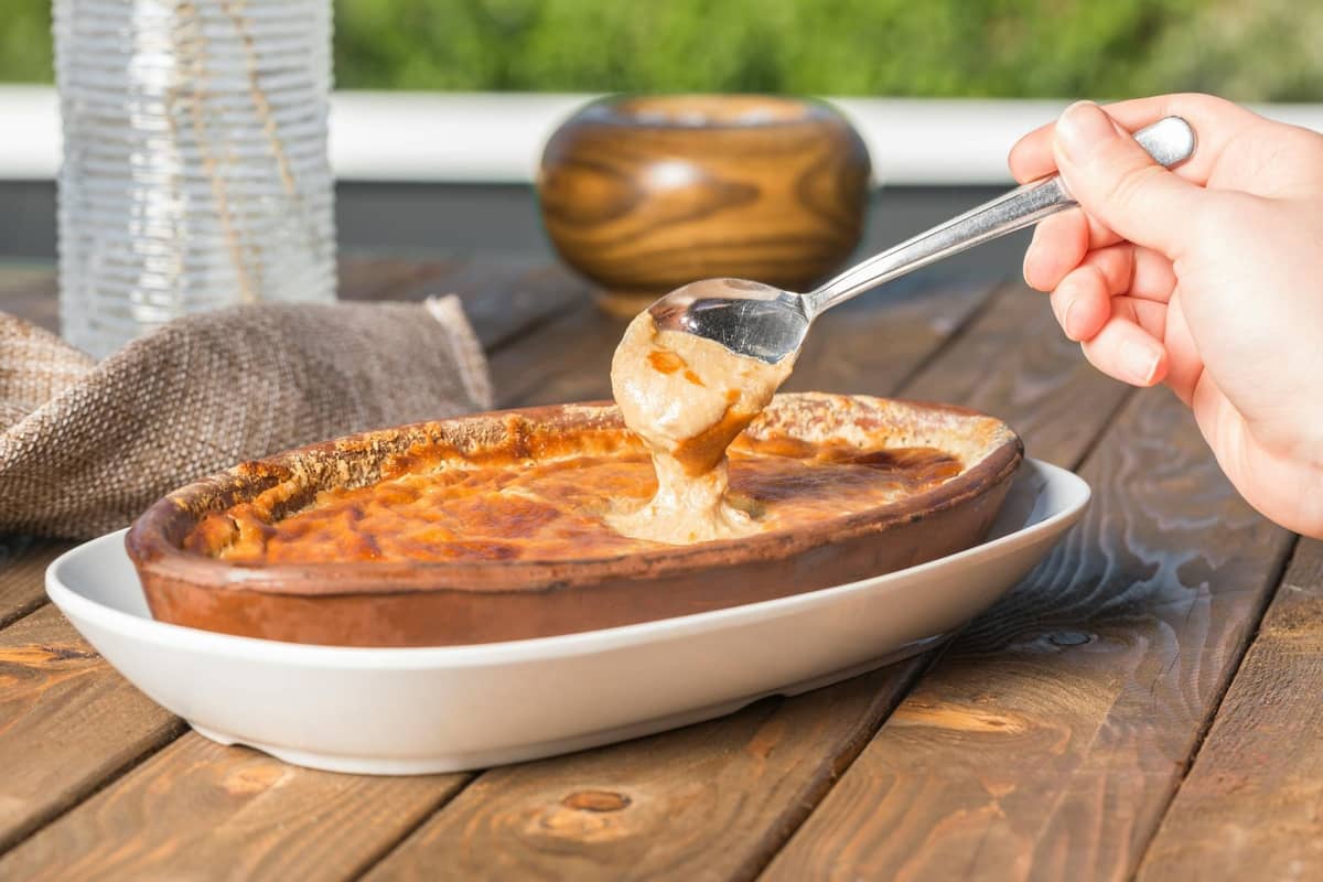Hand scooping creamy yellow dip from oval clay dish, likely Santorini fava, on a wooden table outdoors.