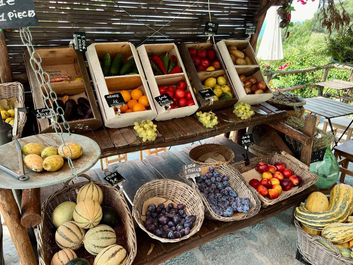 Outdoor market stand in Santorini with baskets and boxes of seasonal local produce including melons, plums, tomatoes, cucumbers, and grapes.