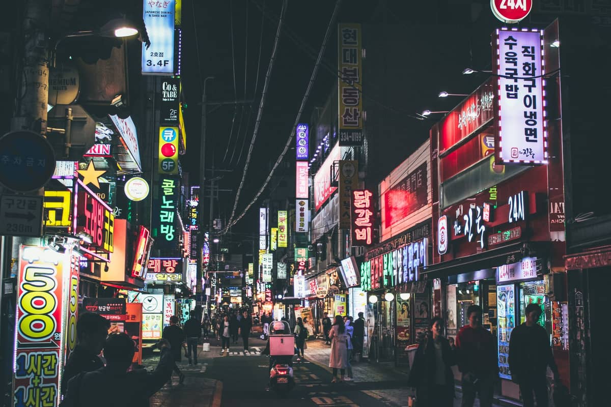 Movimentada rua de Seul à noite repleta de letreiros de néon de restaurantes e bares, pessoas caminhando e jantando, vibrante atmosfera noturna.