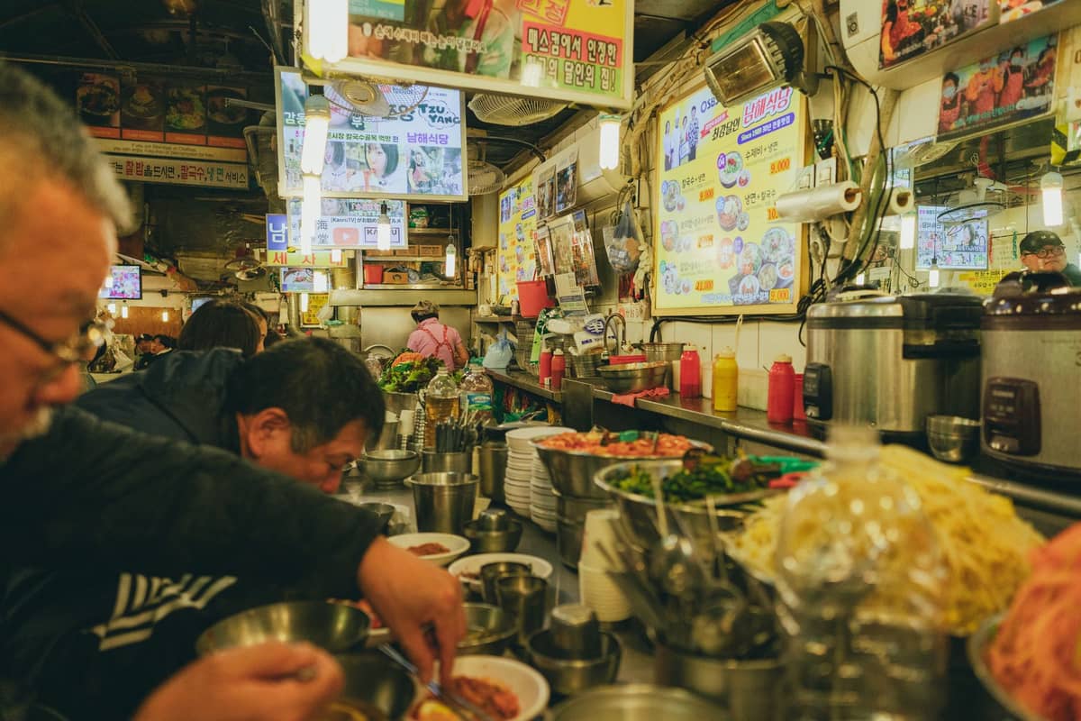 Barraca de comida em um movimentado mercado de Seul com pessoas comendo, cercada de pratos, ingredientes e coloridas placas de menu coreanas nas paredes.