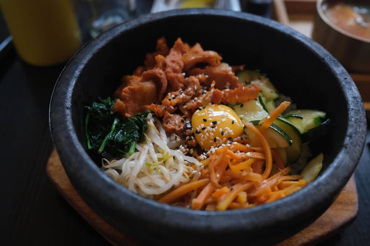 Um close-up de dolsot bibimbap em uma tigela de pedra quente com vegetais variados, carne fatiada, gema de ovo crua e sementes de gergelim.