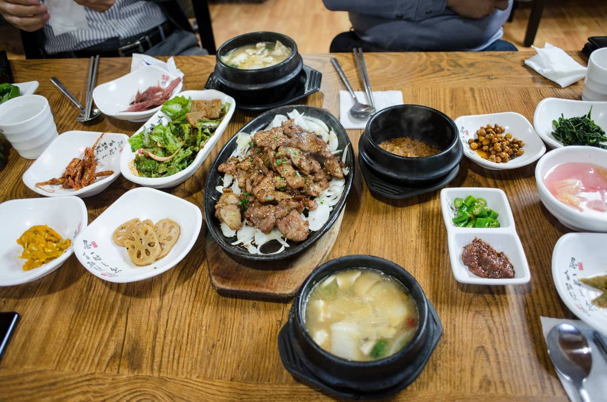 Vista ampla de uma mesa de refeição coreana em Seul com carne grelhada como prato principal e variados banchan em pequenas tigelas, com uma disposição de jantar comunal.
