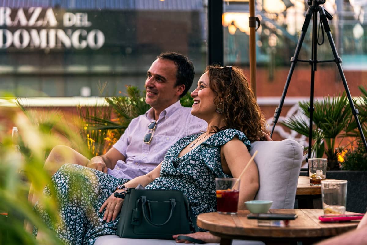 สองคนกำลังดื่มเครื่องดื่มบนระเบียงที่แดดสดใสด้านหน้าป้าย 'PLAZA DEL DOMINGO' โดยมีแก้วต่างๆ และต้นไม้ให้เห็น