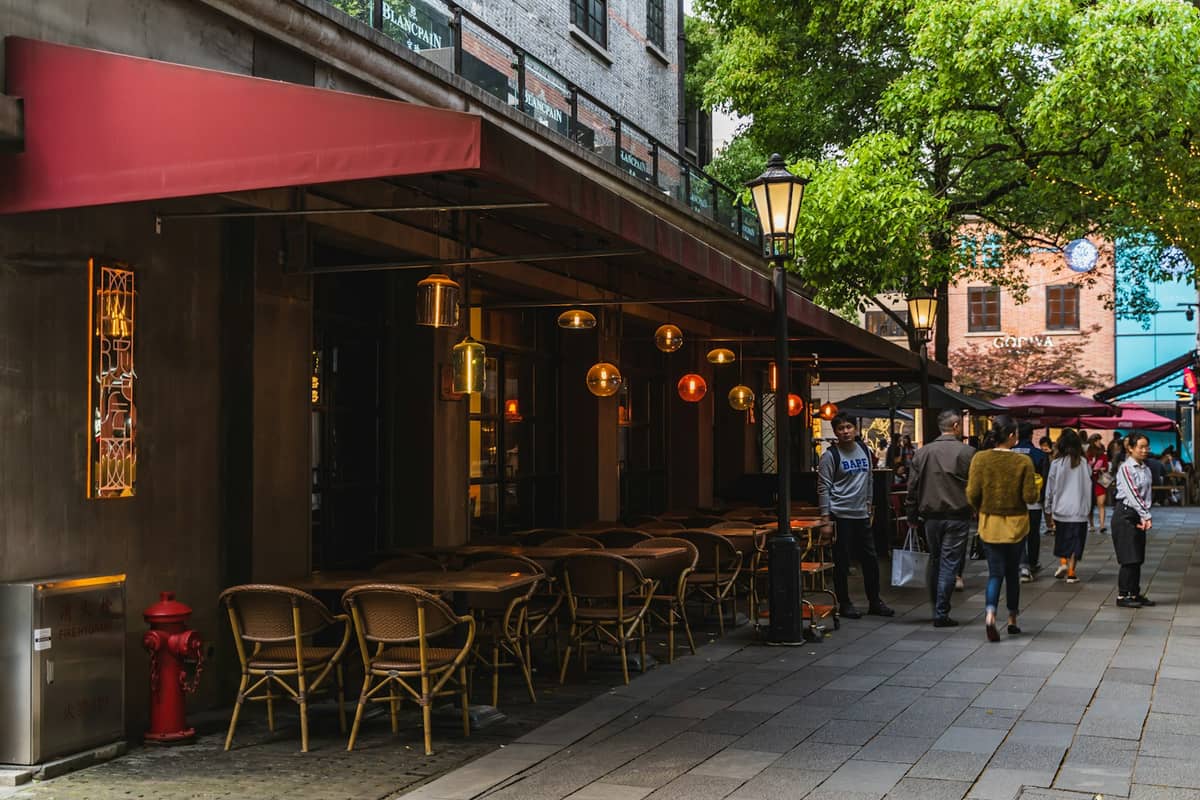 Outdoor dining area of a restaurant with red awning on a leafy pedestrian street in Shanghai, people walking along the sidewalk