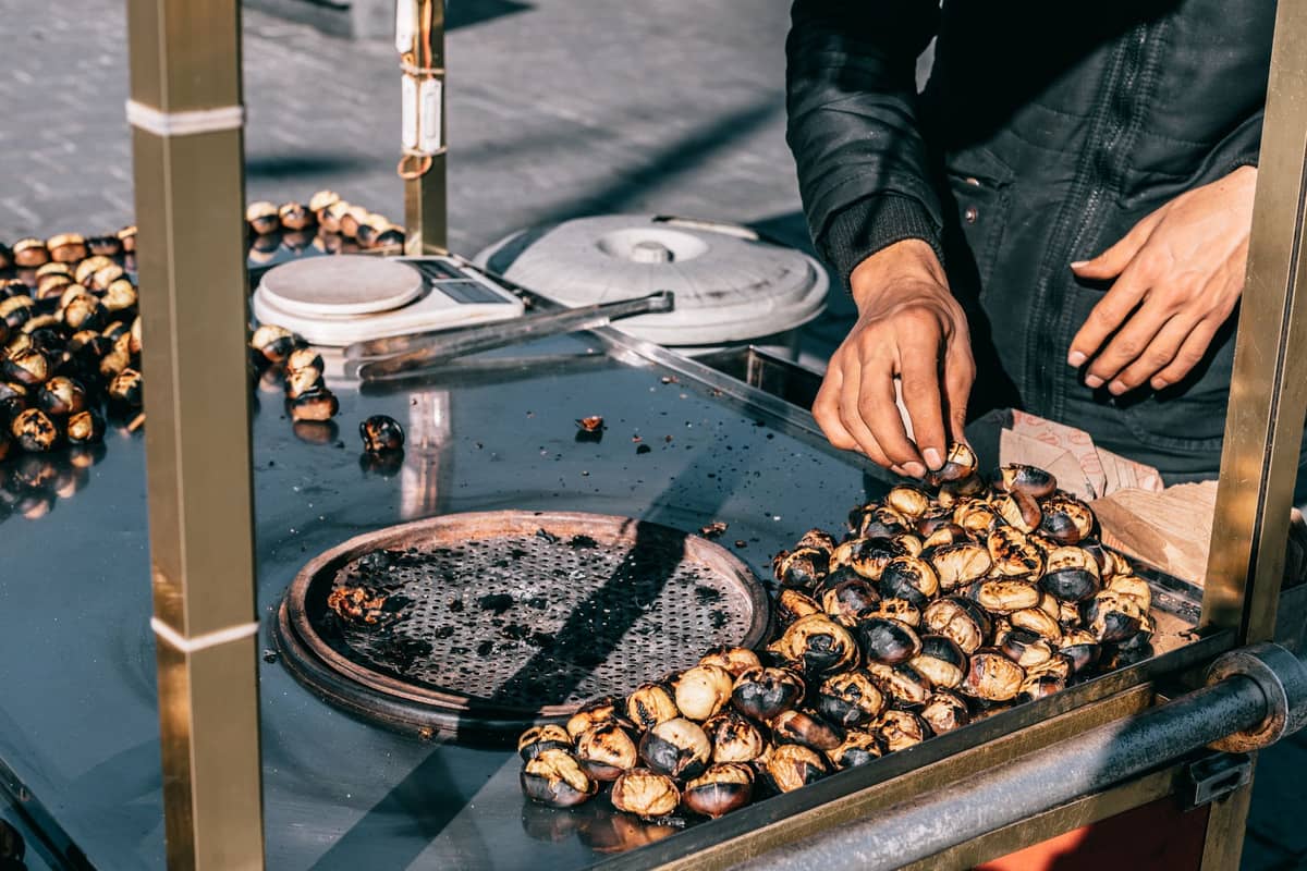 Street vendor roasting and selling chestnuts at a metal cart, with hands visible and many freshly roasted chestnuts in view.