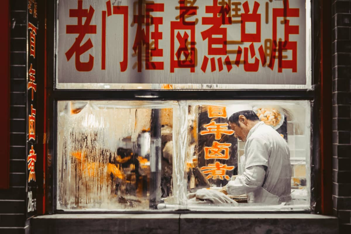 View through a steamed restaurant window showing a chef preparing food at a local Chinese eatery with large red Chinese characters above.