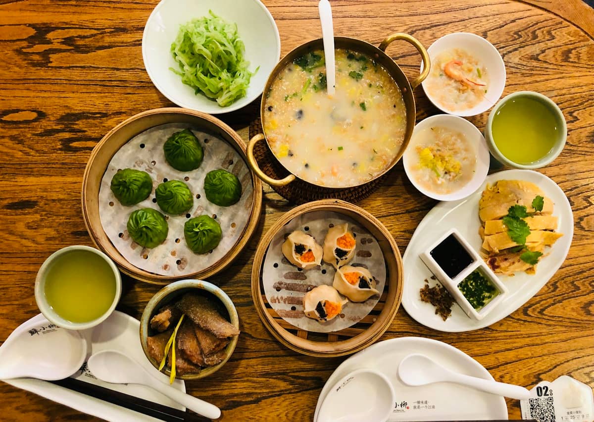 Ein Holztisch mit einer Auswahl klassischer Shanghaier Gerichte: Dumplings, Hähnchen, Suppe, Gemüse und Tee in einem Restaurant.