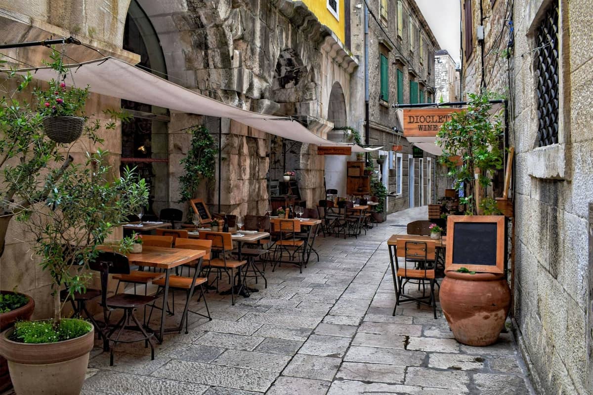 Rue étroite en pierre à Split avec des tables de restaurant en terrasse et des enseignes, ruelle atmosphérique entourée de bâtiments historiques.