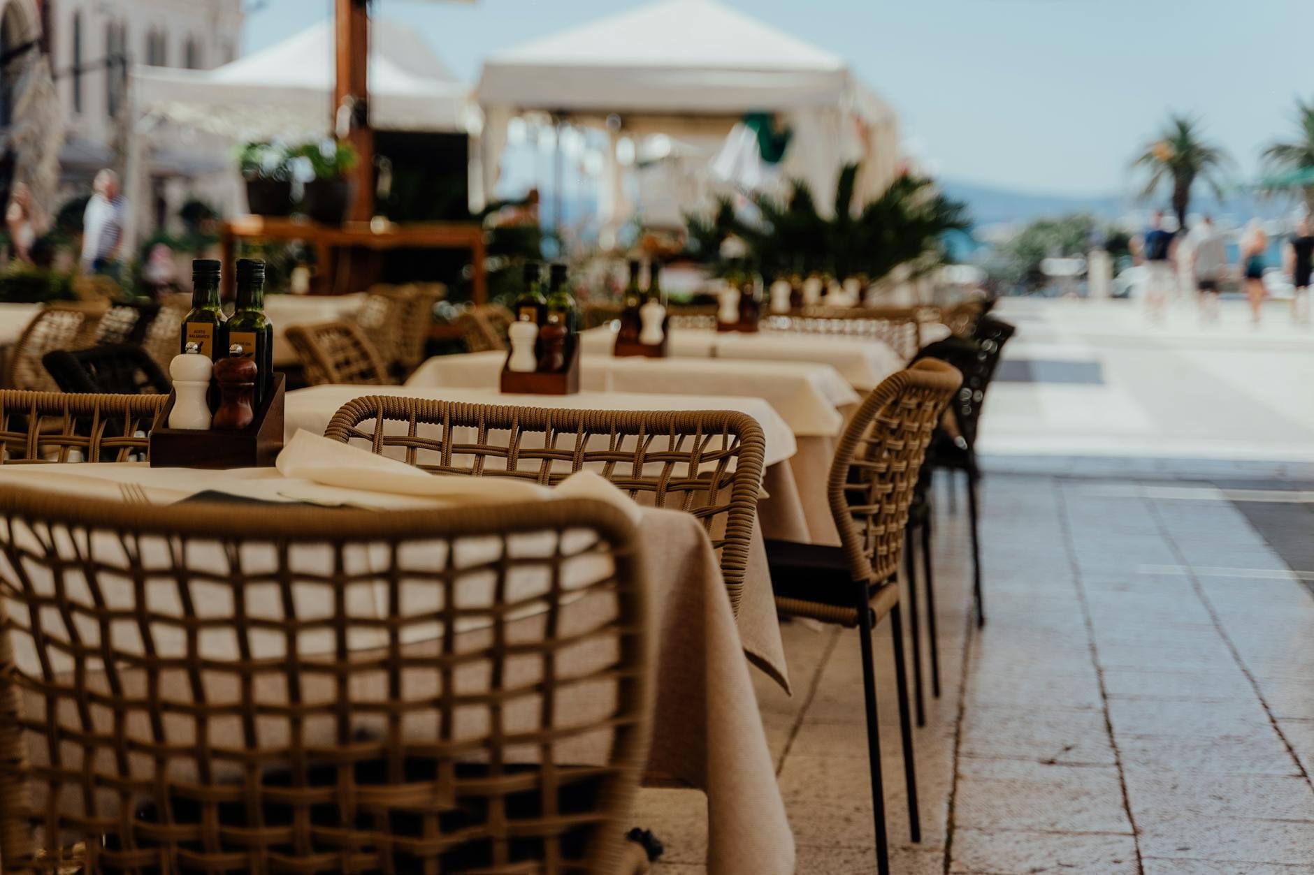 Mesas de restaurante à beira-mar com azeite e temperos na orla Riva de Split, atmosfera acolhedora com palmeiras e vista para o Adriático ao fundo.