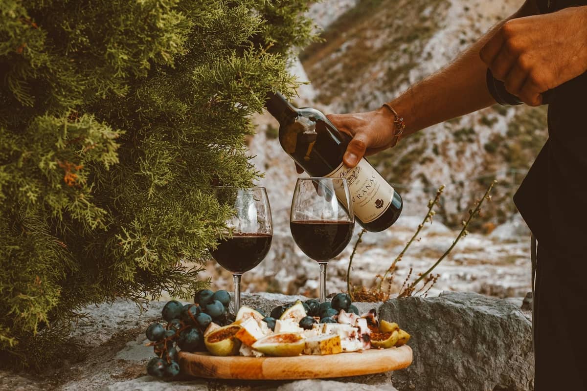Une personne verse du vin rouge dans deux verres à côté d'un plateau de fromage, de raisins et de figues sur une surface en pierre rustique en plein air.
