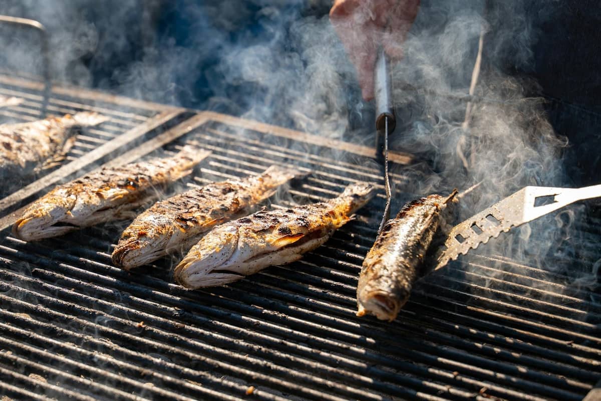 Plusieurs poissons entiers grillant sur un barbecue au charbon de bois avec de la fumée visible et une main retournant un poisson sur le gril.