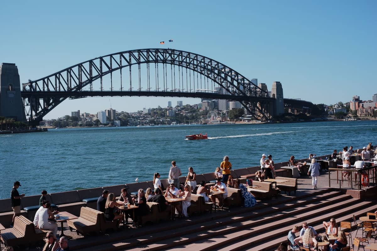 Zatłoczony taras restauracyjny nad wodą, goście jedzą i piją, w tle Most Harbour Bridge w słoneczny dzień.