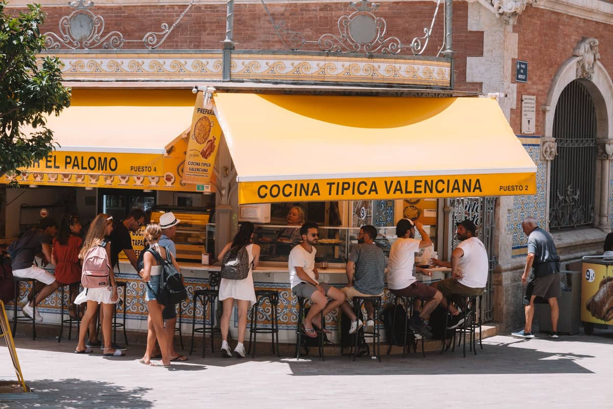 Lockere Menschenmenge an einem Außentresen unter gelbem Vordach mit der Aufschrift Cocina Tipica Valenciana, die den alltäglichen Essrhythmus in Valencia, Spanien, widerspiegelt.