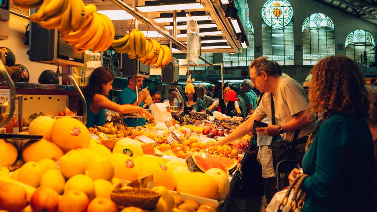 Visitatori al Mercado Central di Valencia tra bancarelle di frutta e verdura, con banane appese e le vetrate colorate sullo sfondo.