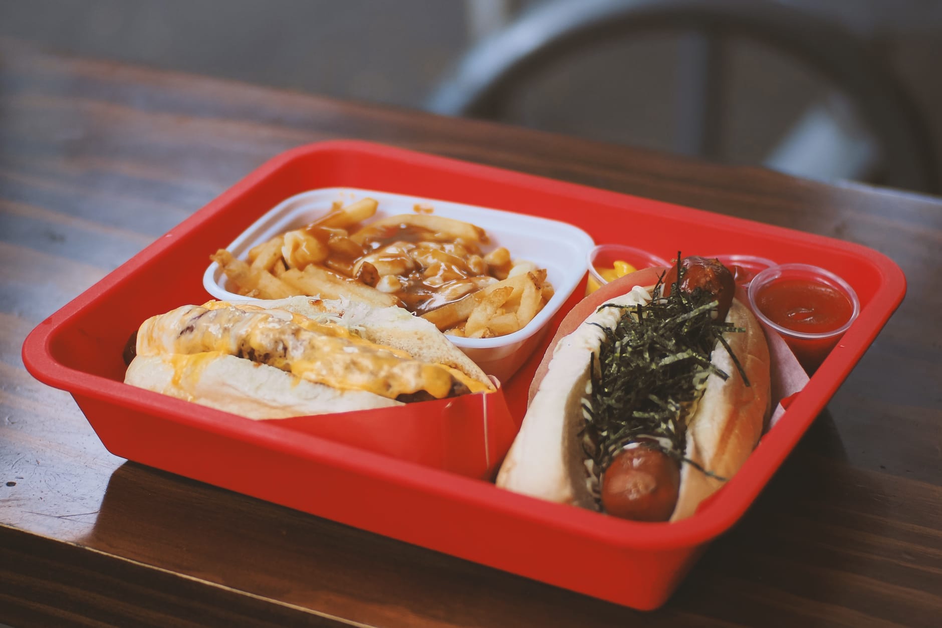 Uma bandeja vermelha com um cachorro-quente estilo japonês, um cachorro-quente com queijo e poutine com fritas, servidos em uma mesa de madeira em Vancouver.