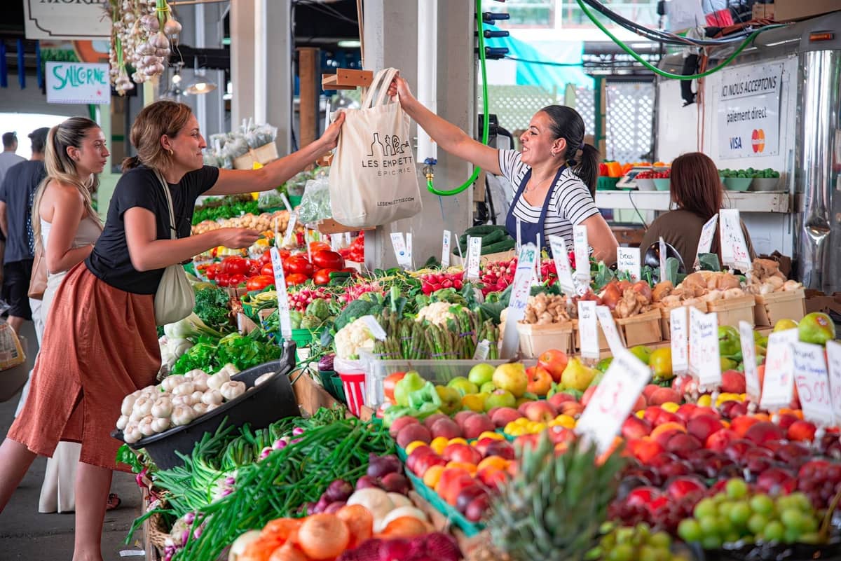 Lebhafte Szene eines überdachten Bauernmarkts in Vancouver mit Menschen, die frische Produkte kaufen, darunter Pfirsiche, Kirschen, Grüngemüse und mehr im Sommer.