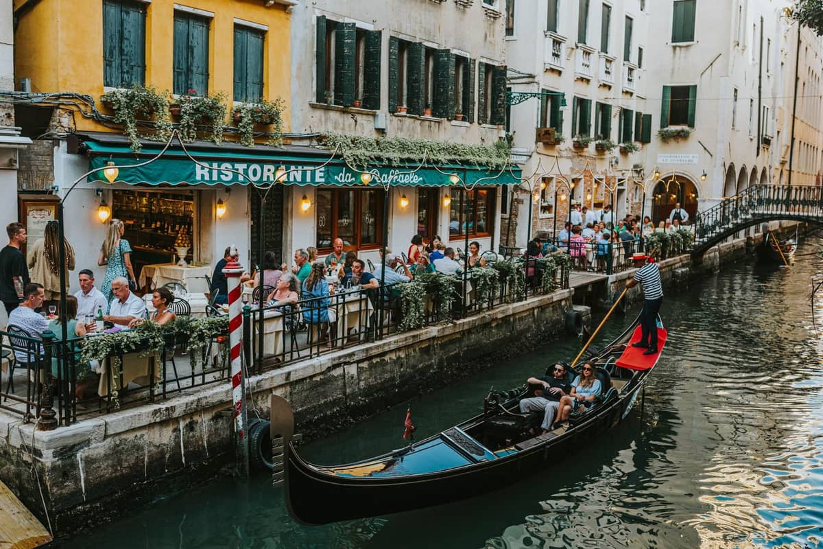 Belebtes venezianisches Kanalrestaurant mit Gästen auf der Außenterrasse, während eine Gondel am Wasser vorbeifährt.