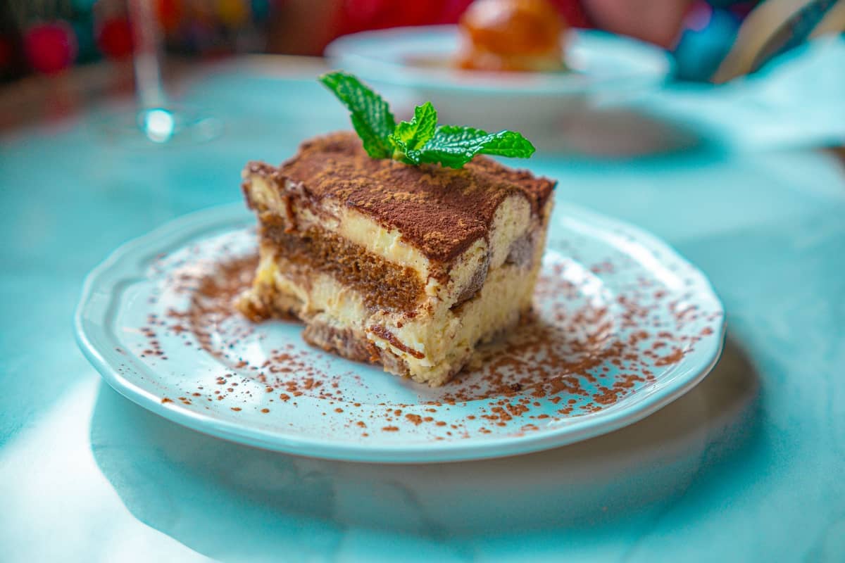 Ein klassisches Stück Tiramisù auf einem weißen Teller, mit Kakaopulver bestäubt und mit frischer Minze garniert, auf einem blauen Tisch.