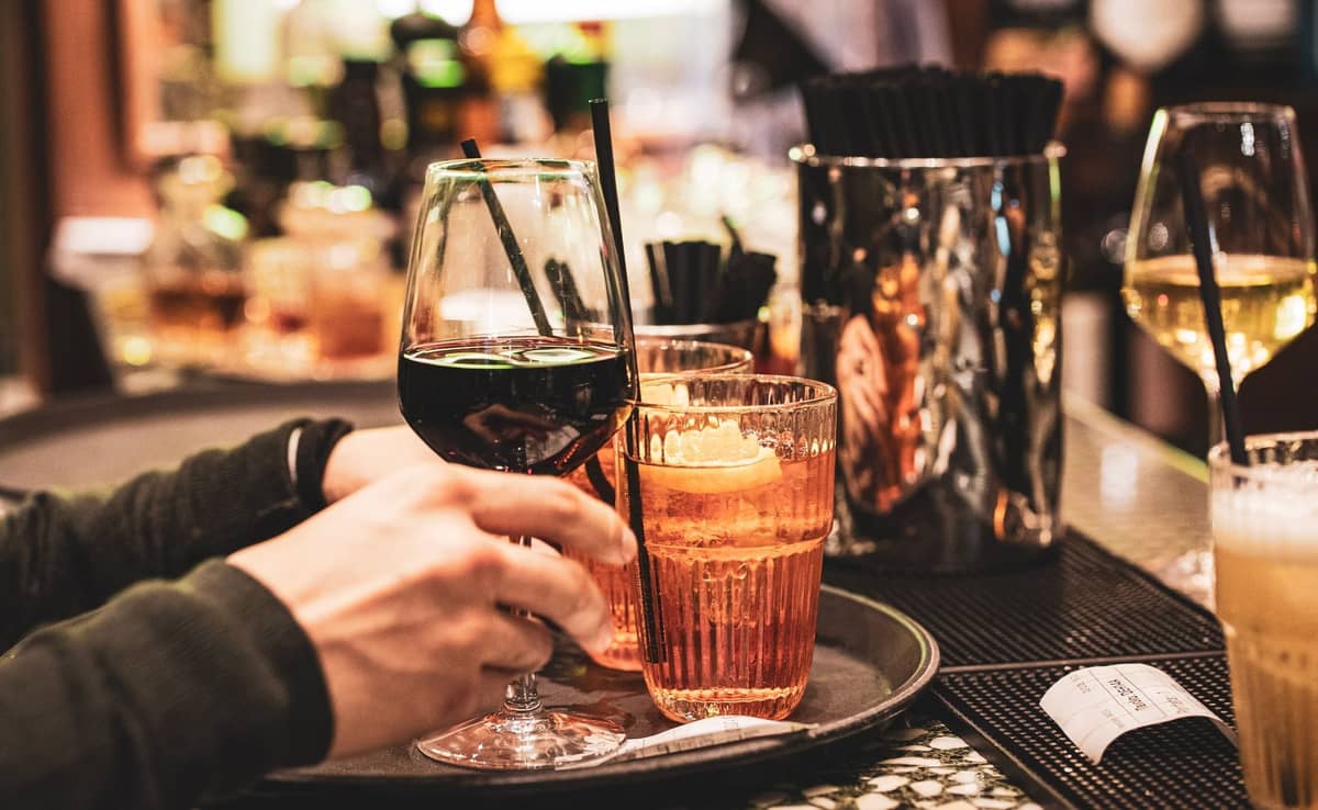 Nahaufnahme eines Bartabletts mit Gläsern Rotwein, Aperol Spritz und anderen Getränken, die in einem lebhaften Café serviert werden.