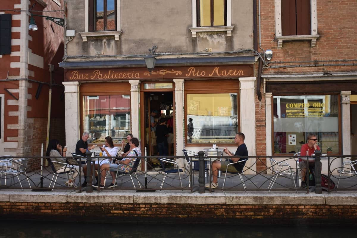 Menschen, die draußen an einer traditionellen venezianischen Bar entlang eines Kanals sitzen und in einem typischen Bacaro Getränke und Snacks genießen.