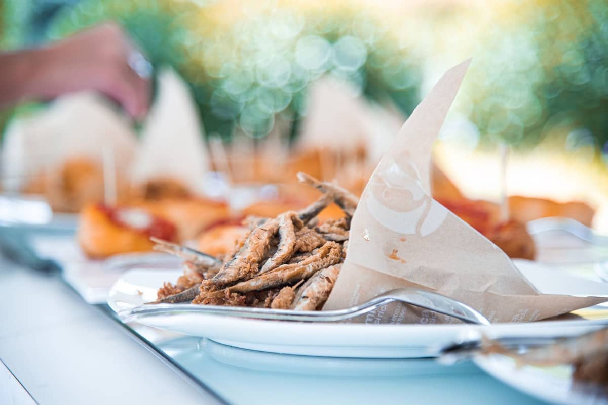 Teller mit frisch gebratenen kleinen Fischen in Papiertüten, typisch für die venezianischen Cicchetti, auf einem hellen Tisch im Freien.