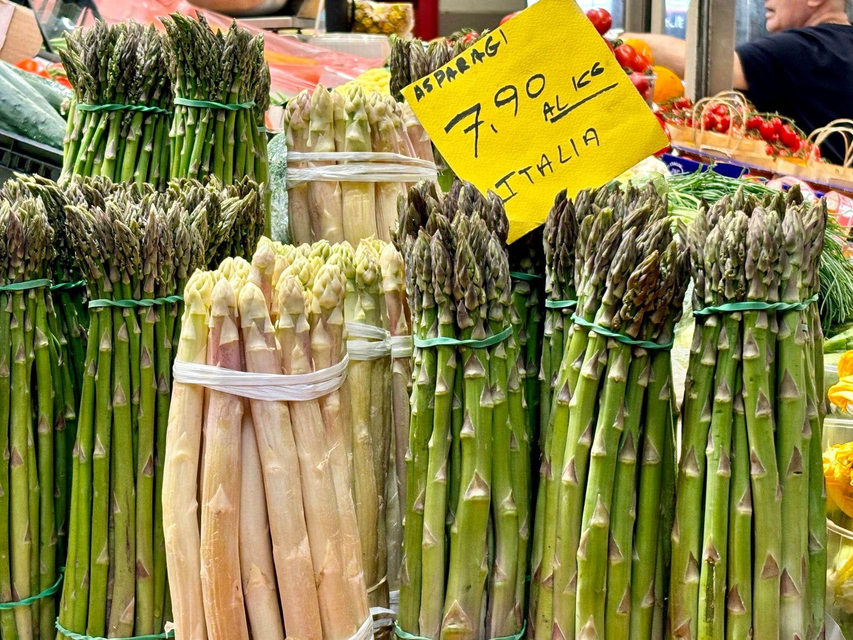 Bündel grüner und weißer Spargel zum Verkauf an einem Frischmarktstand, mit einem handgeschriebenen Preisschild.