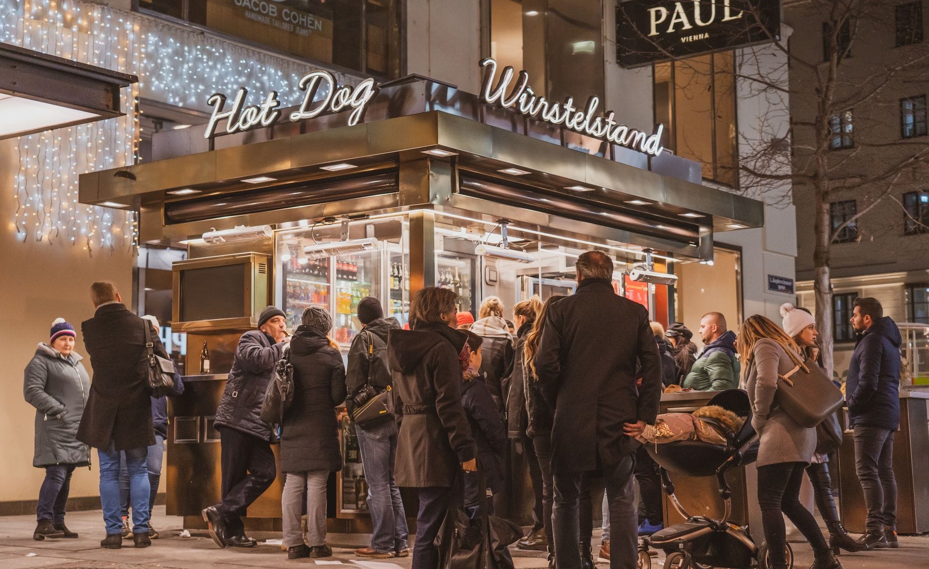 Ein belebter Wiener Würstelstand bei Nacht, um den sich Menschen versammelt haben und typisches Wiener Streetfood genießen.