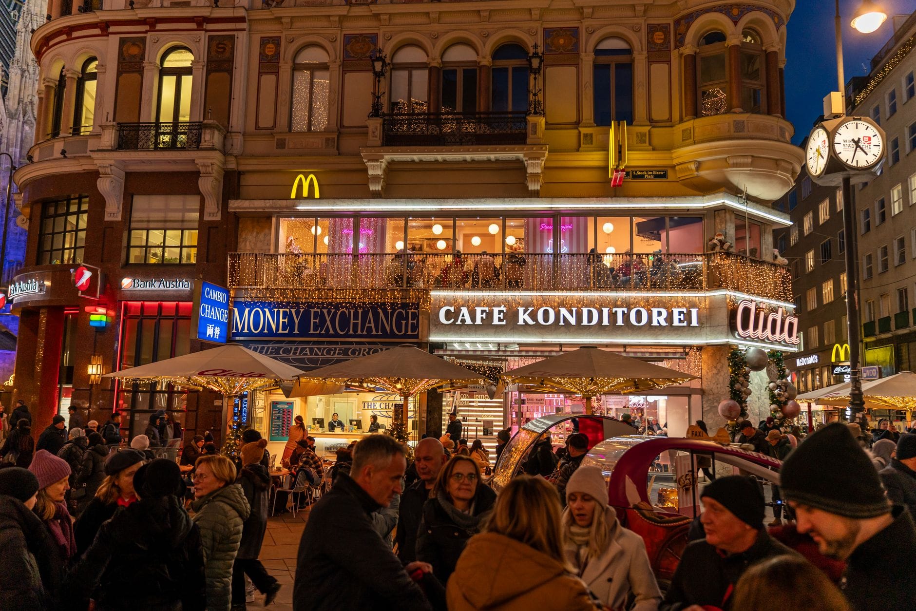 Menschenmenge vor einem historischen Café Konditorei in Wien, nachts beleuchtet mit dekorativen Lichtern und klassischer Architektur.
