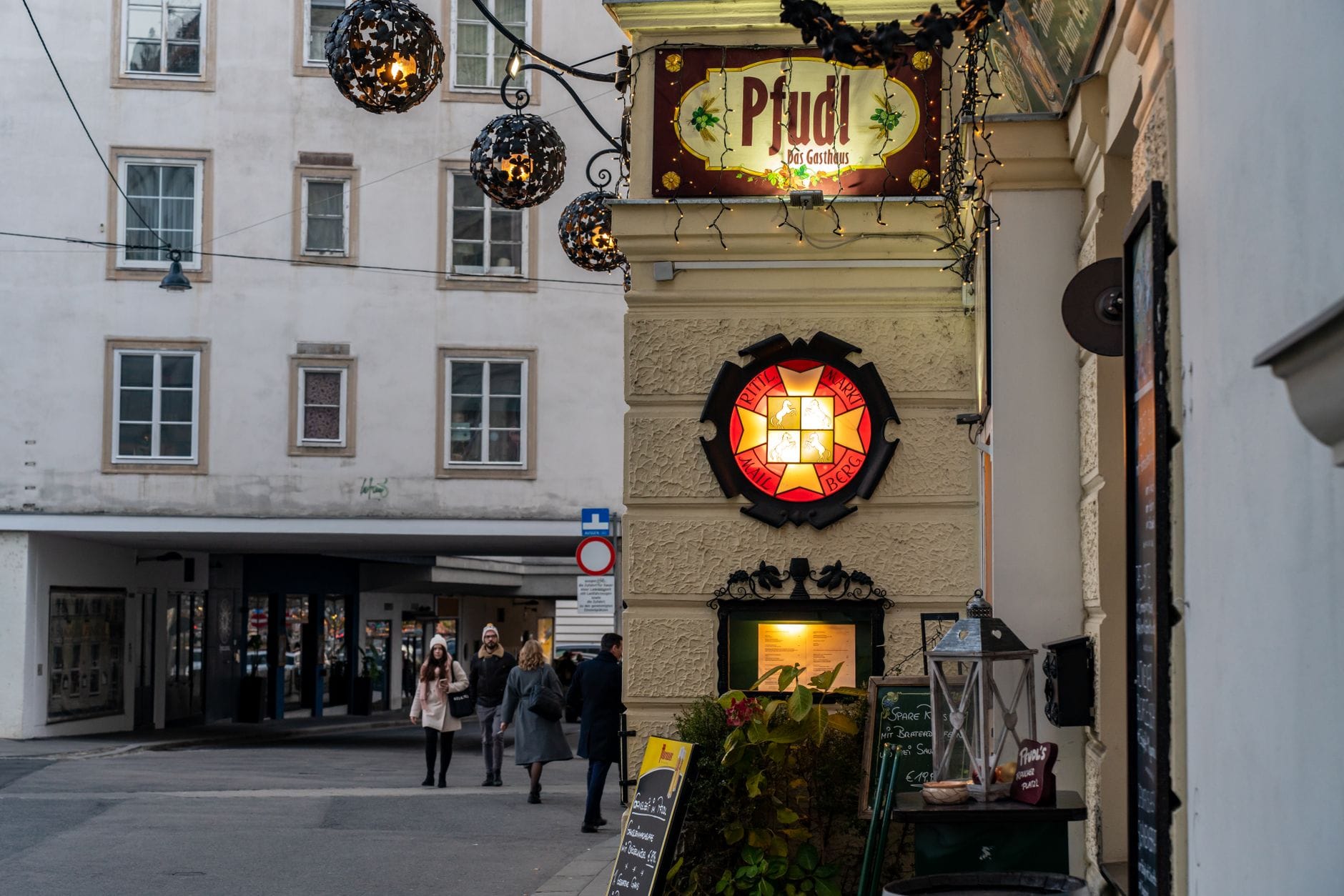 Straßenansicht des Eingangs eines traditionellen Wiener Restaurants mit Beschilderung, dekorativen Lichtern und vorbeigehenden Menschen.