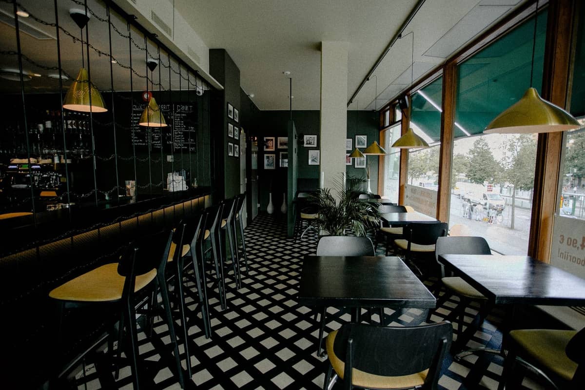Intérieur de bistrot moderne à Varsovie avec de grandes fenêtres, un sol carrelé noir et blanc, des tables et chaises noires et des suspensions dorées.