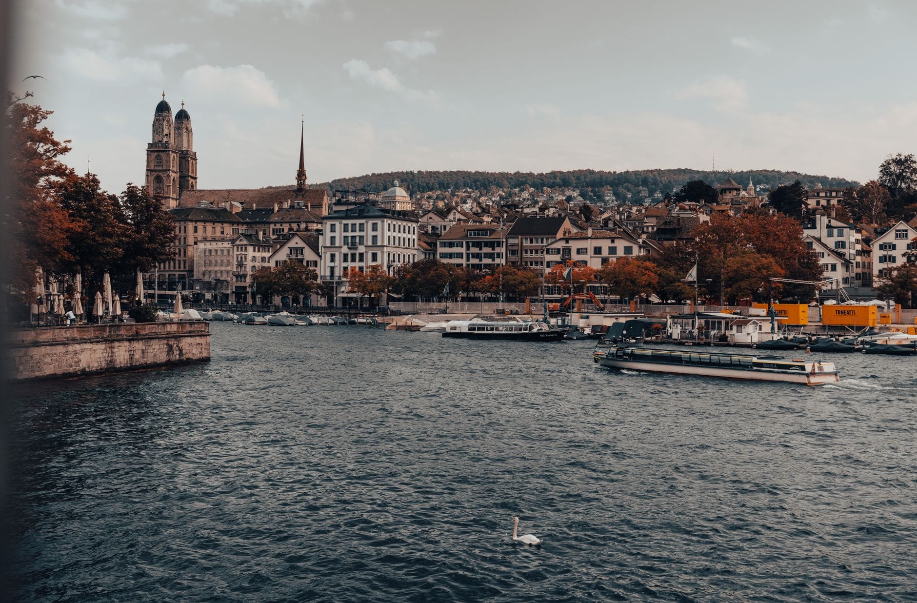 วิวกว้างใจกลางเมืองซูริกริมแม่น้ำ Limmat เห็นแถวอาคารริมน้ำ หอคอยโบสถ์ และเรือในน้ำ ใต้ท้องฟ้ามีเมฆปกคลุม
