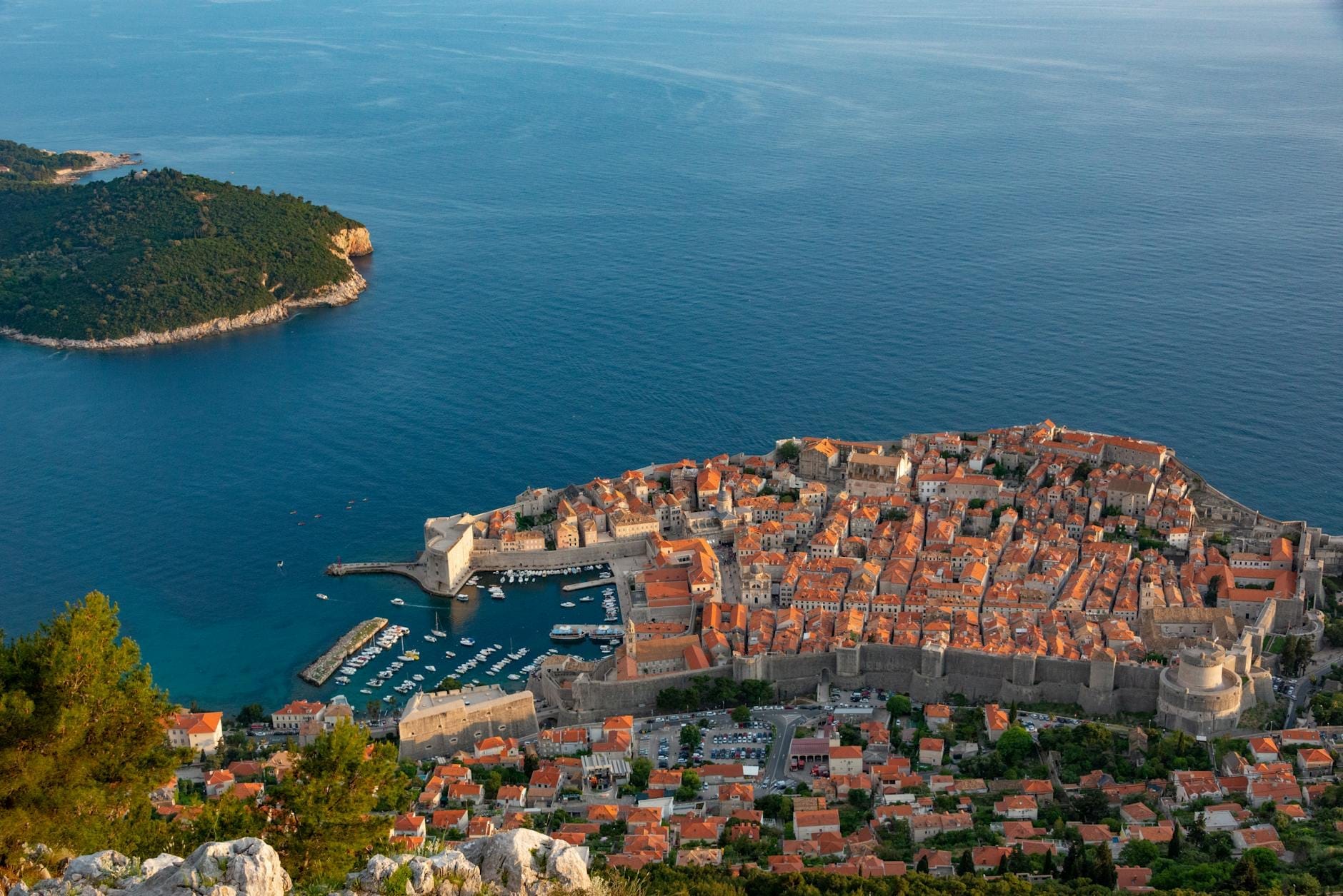 Vista aérea del casco antiguo de Dubrovnik en tierra firme con la isla de Lokrum cerca, separados por el mar azul bajo un cielo despejado.