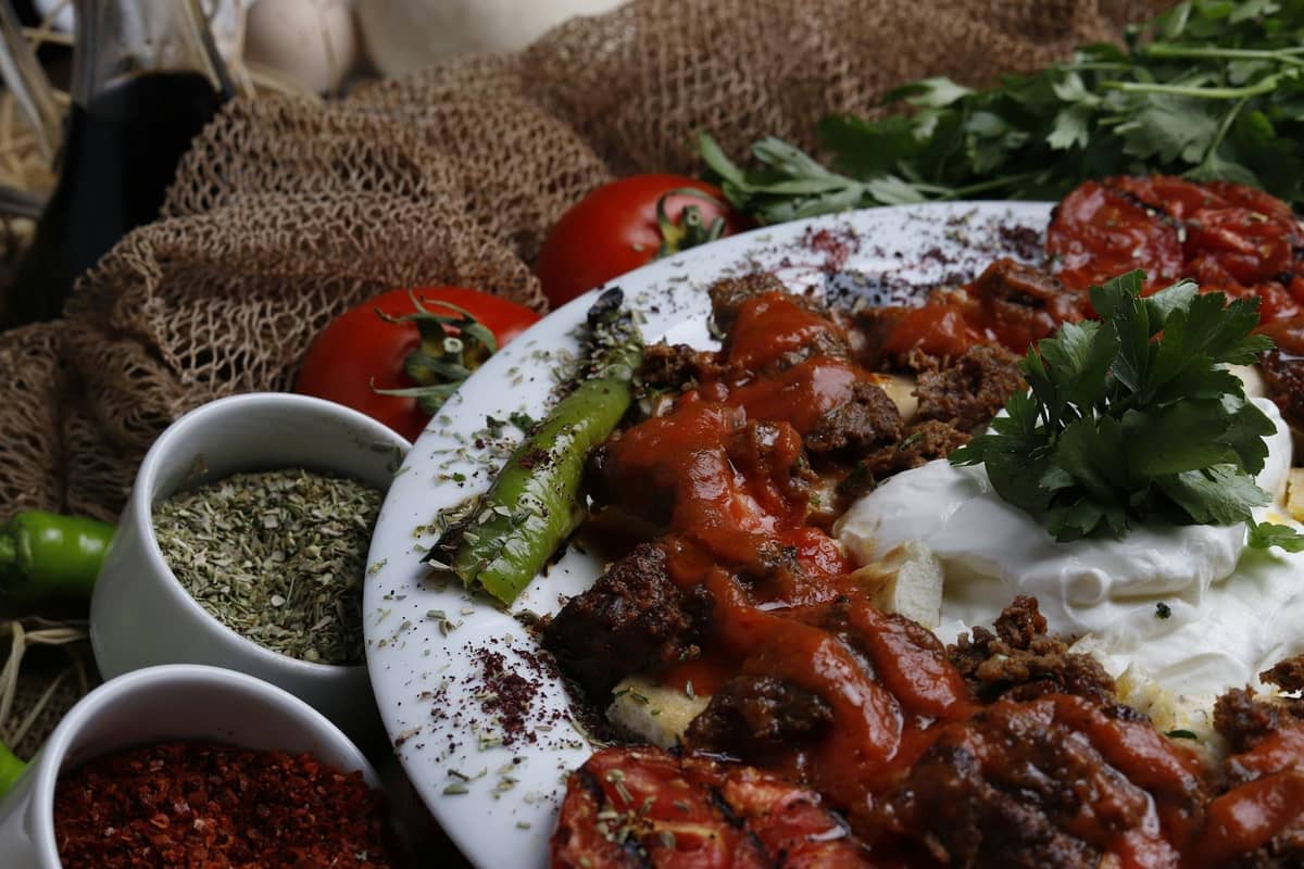 Close-up of a Turkish dish with grilled meat, tomato, pepper, yogurt, and herbs on a plate surrounded by spices and fresh vegetables.