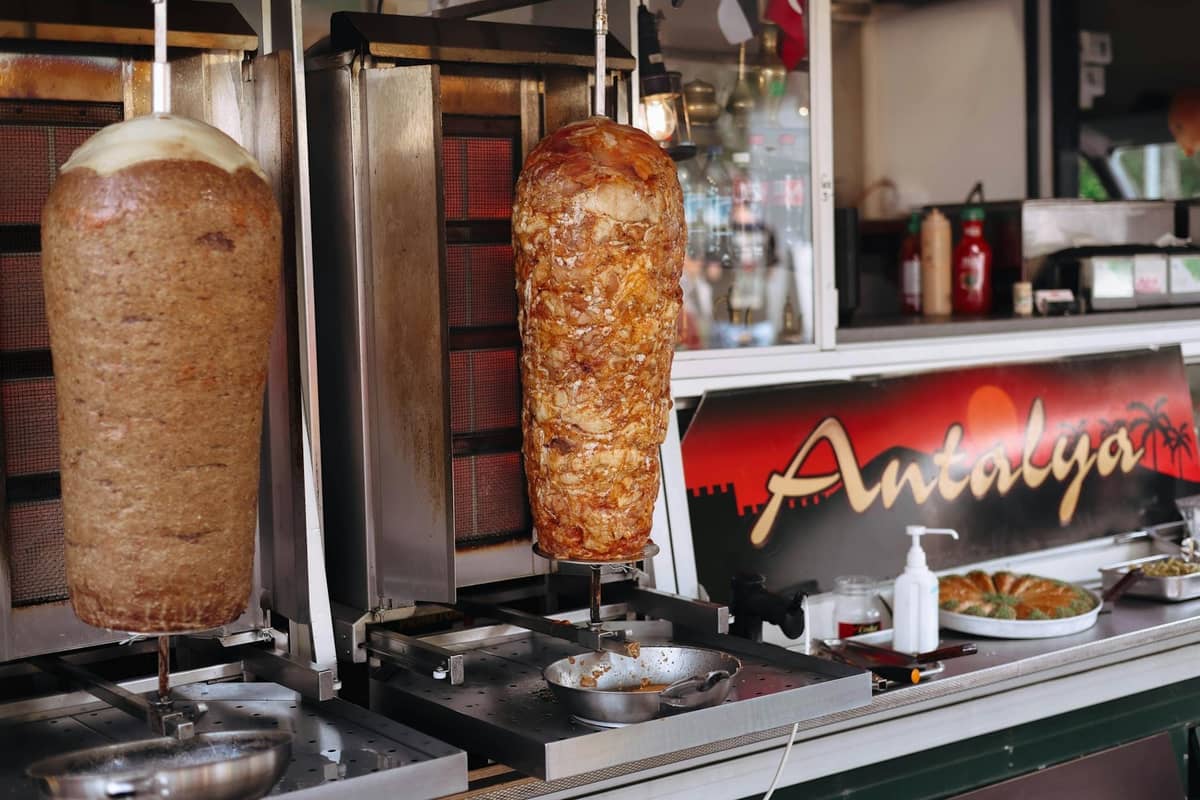 Doner kebab meat rotating on spits in a street food stall, with a sign that says 'Antalya' in the background.