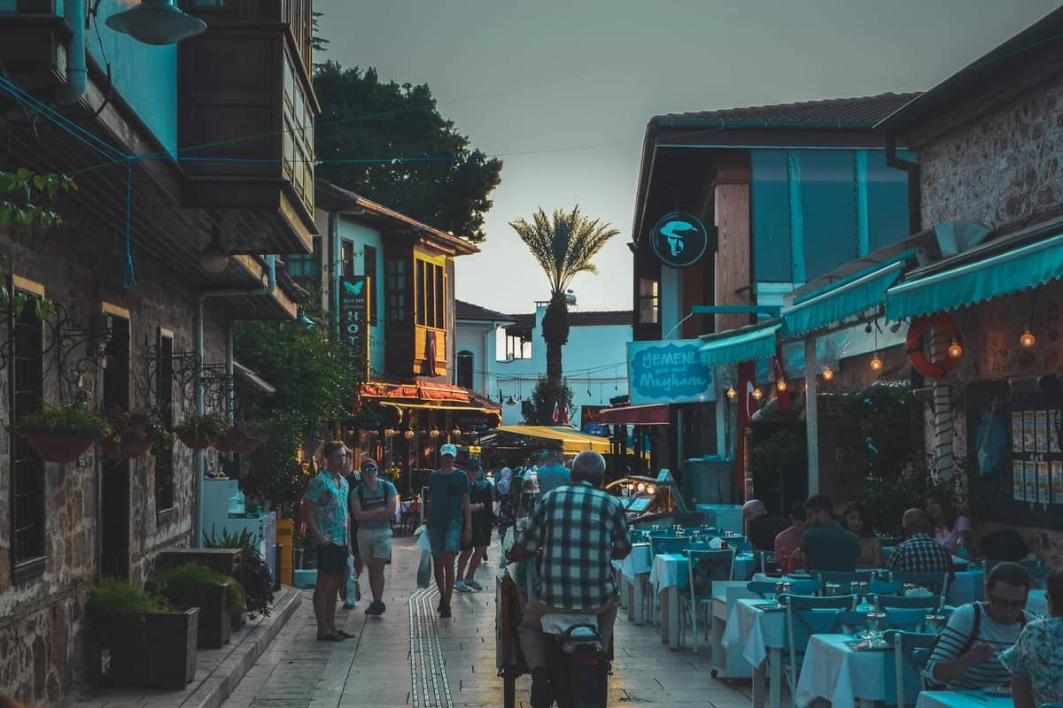 Evening view of a lively street in Kaleiçi, Antalya, with outdoor restaurant seating, historic buildings, and people strolling.