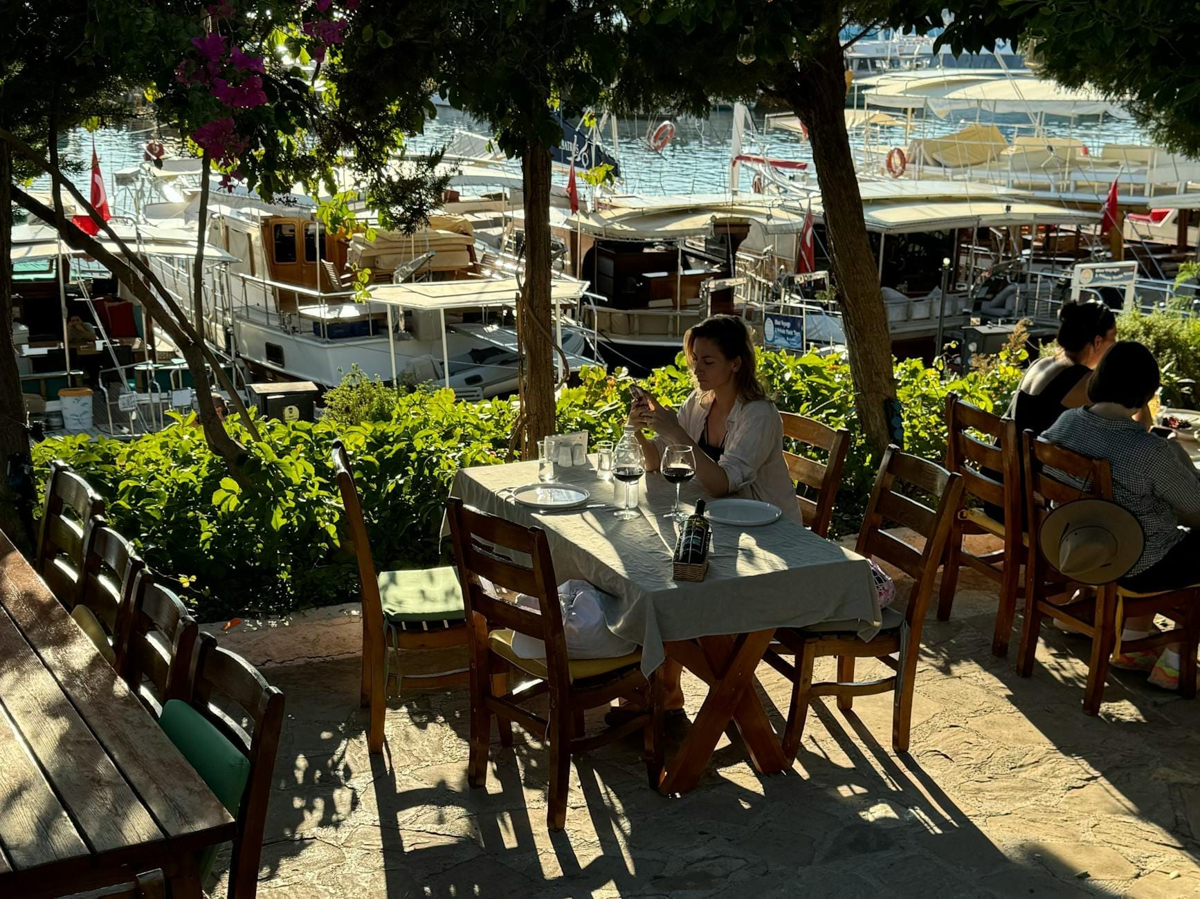 Mesas de restaurante ao ar livre com vista para a marina de Antalya, com mesas de madeira, sombra de árvores, pessoas jantando e iates brancos atracados ao fundo.