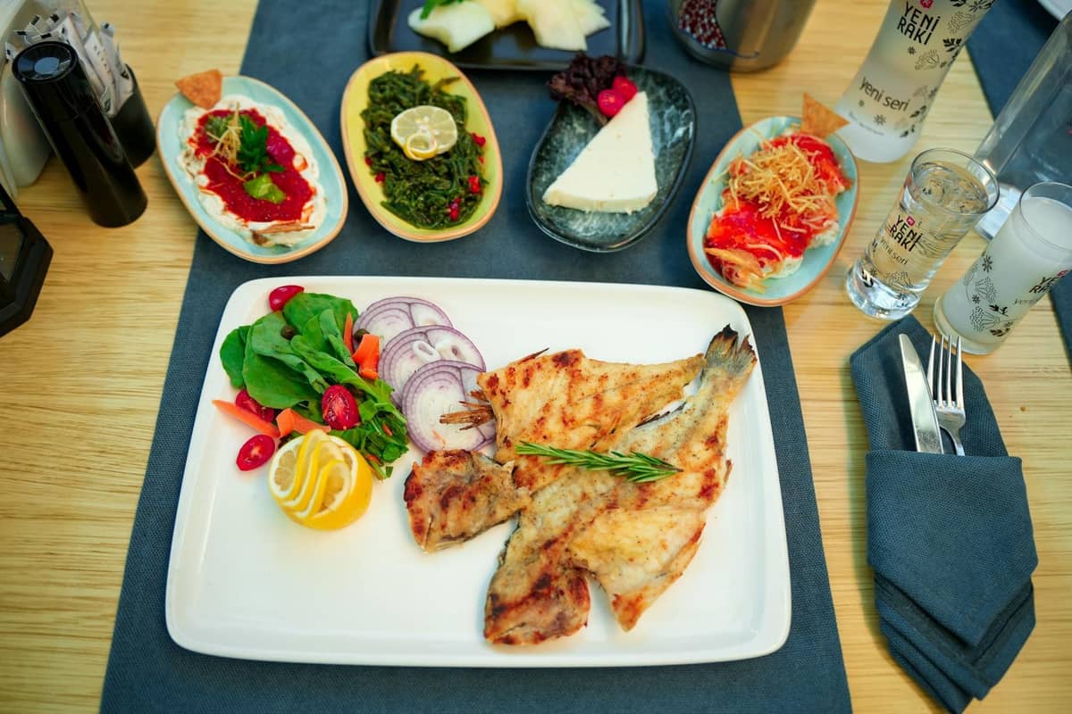 A well-lit restaurant table with grilled fish, fresh salad, Turkish mezes, and raki, showcasing Mediterranean cuisine associated with Antalya’s food scene.